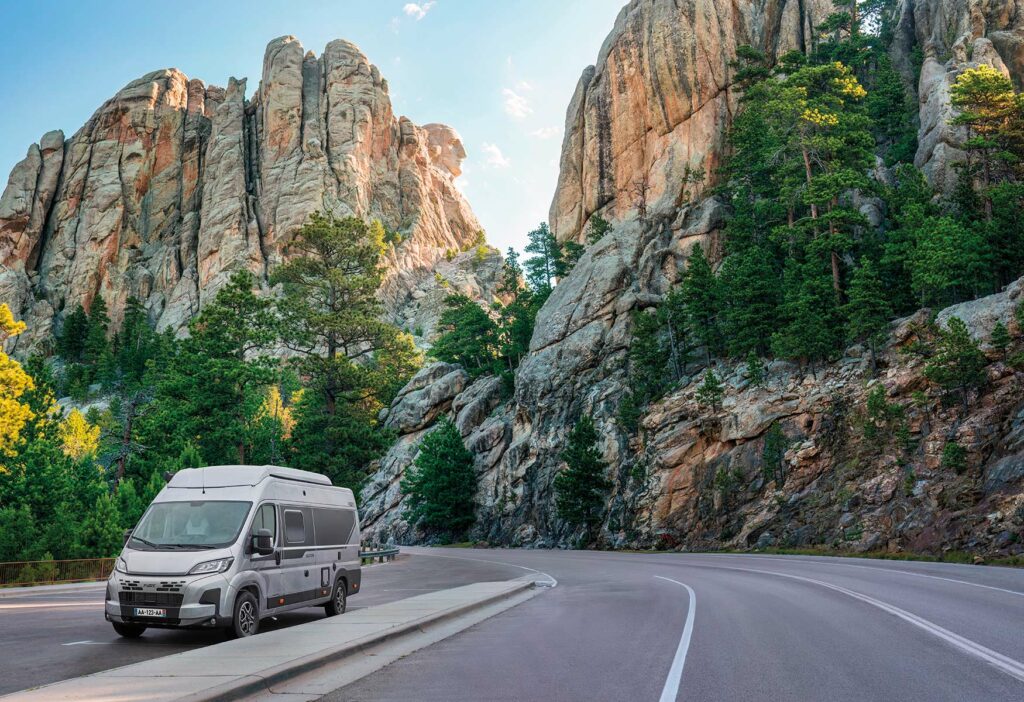 Fourgon Pilote gris avec stickers noirs roulant sur une route entourée de hautes falaises et de sapins.